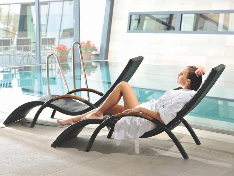 Frau in weißem Bademantel entspannt auf schwarzem Liegestuhl neben Hallenbad in modernem Ambiente.