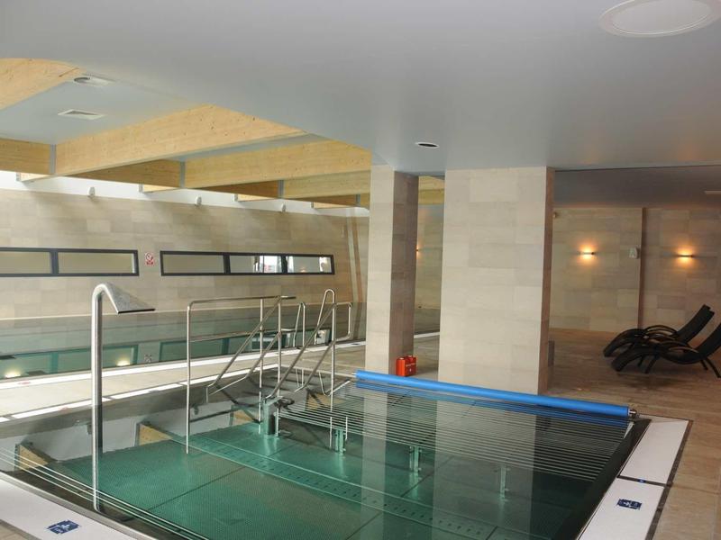 Moderner Innenpool mit Glasgeländer, Holzdeckenbalken und zwei schwarzen Liegestühlen im Hintergrund.