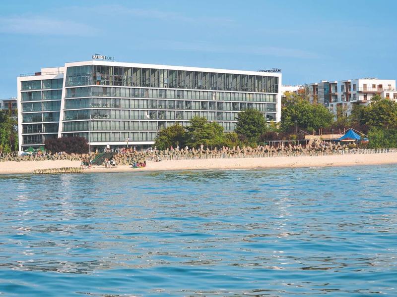 Modernes Gebäude hinter Sandstrand mit Sonnenschirmen und blauem Meer im Vordergrund.
