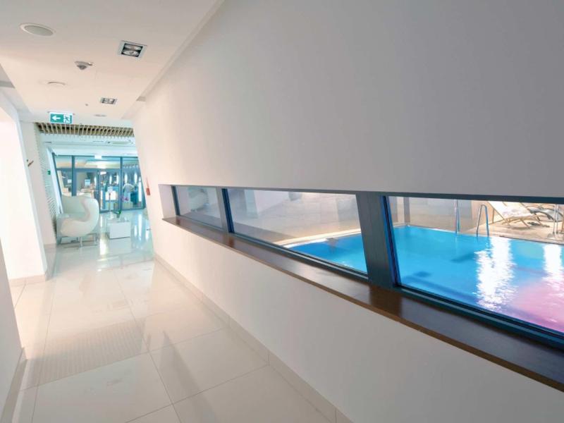 Heller Flur mit weißem Boden, schrägen Fenstern und Blick auf einen Poolbereich.