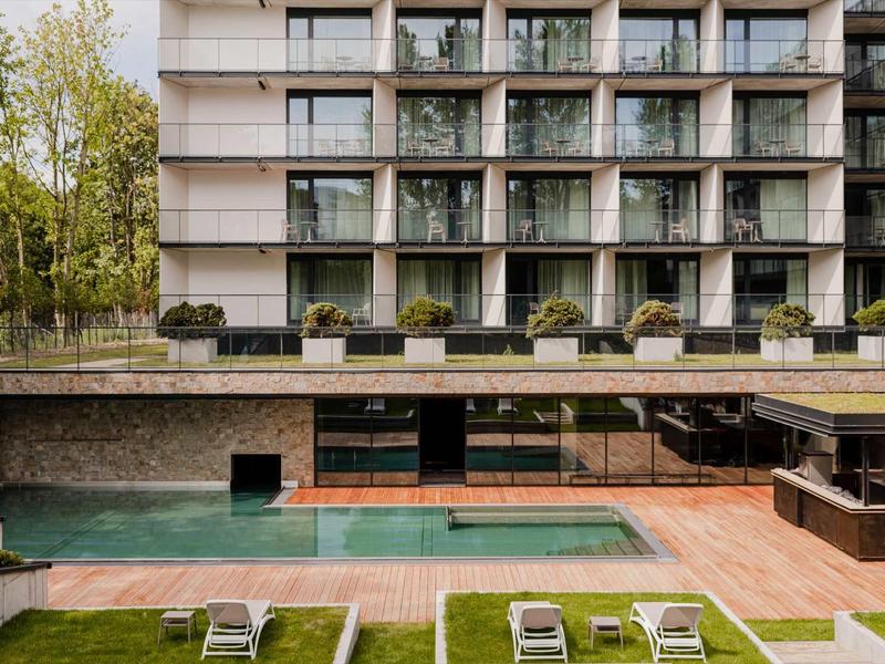 Modernes Gebäude mit großem Pool, Liegestühlen und Grünfläche im Vordergrund, Bäume im Hintergrund.