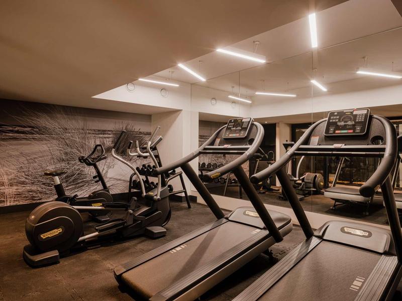 Fitnessraum mit Laufbändern und Crosstrainern, Deckenlichter und Wandspiegel, warme Beleuchtung.