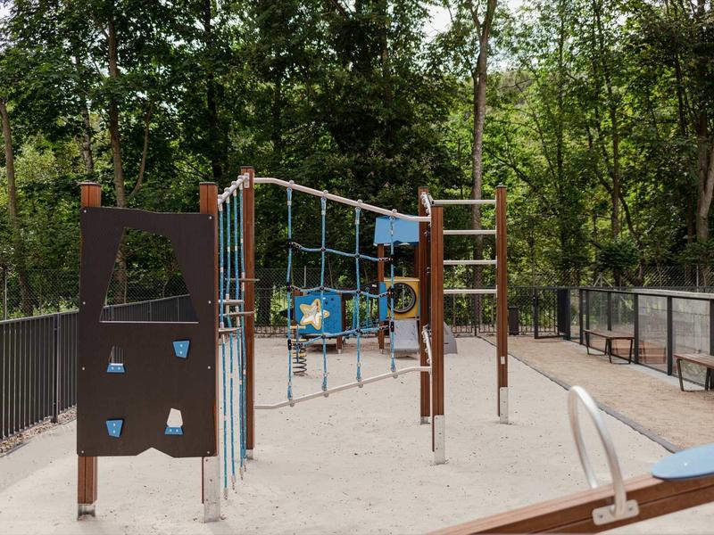 Spielplatz mit Klettergerüst, Netz und Schaukeln, umgeben von Bäumen und Metallzaun.