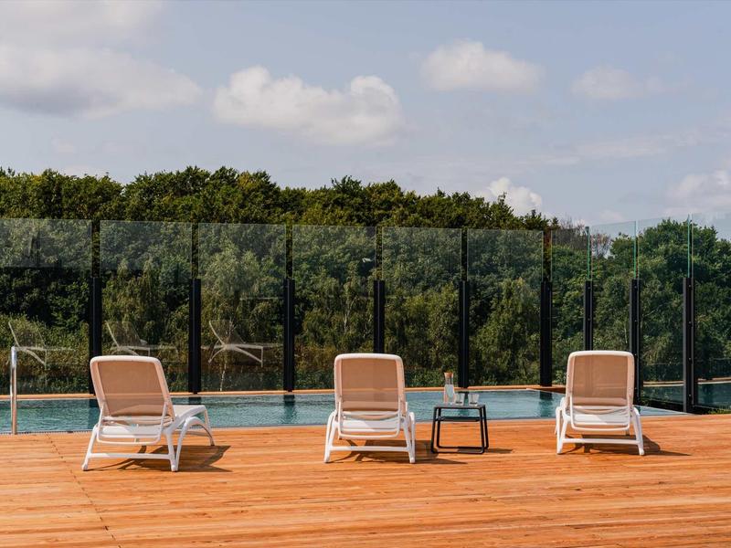 Drei weiße Liegestühle und ein kleiner Tisch auf Holzterrasse vor Pool mit Glastrennwand und Bäumen.
