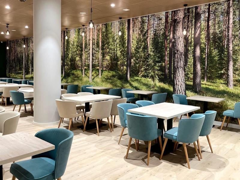Moderner Gastraum mit weißen Tischen, blauen Stühlen und großer Wandgestaltung eines Waldes.