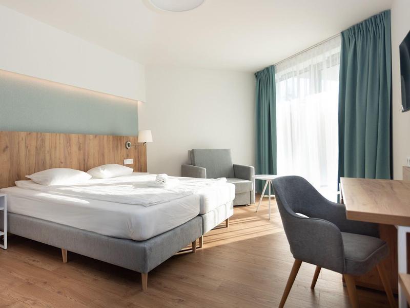 Helles Hotelzimmer mit Doppelbett, grauem Sessel, Schreibtisch und großem Fenster mit grünen Vorhängen.