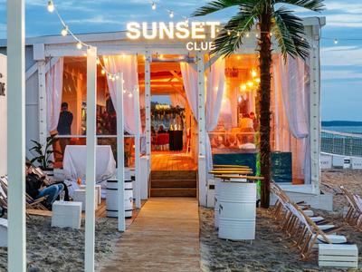 Strandbar met warm licht, witte gordijnen en planten op een zanderig pad bij zonsondergang.