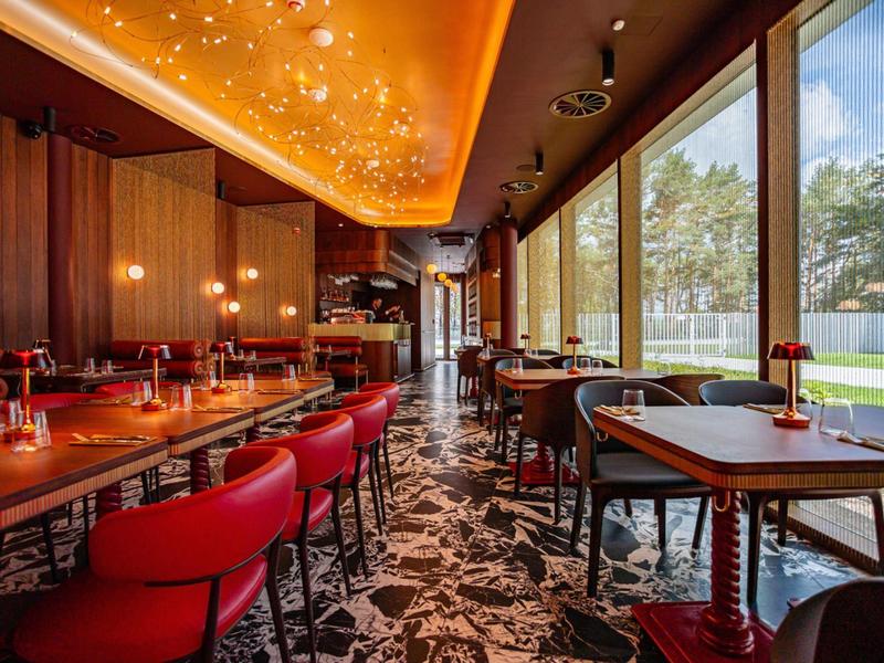 Modern restaurant met rode stoelen, grote ramen en decoratieve plafondverlichting.