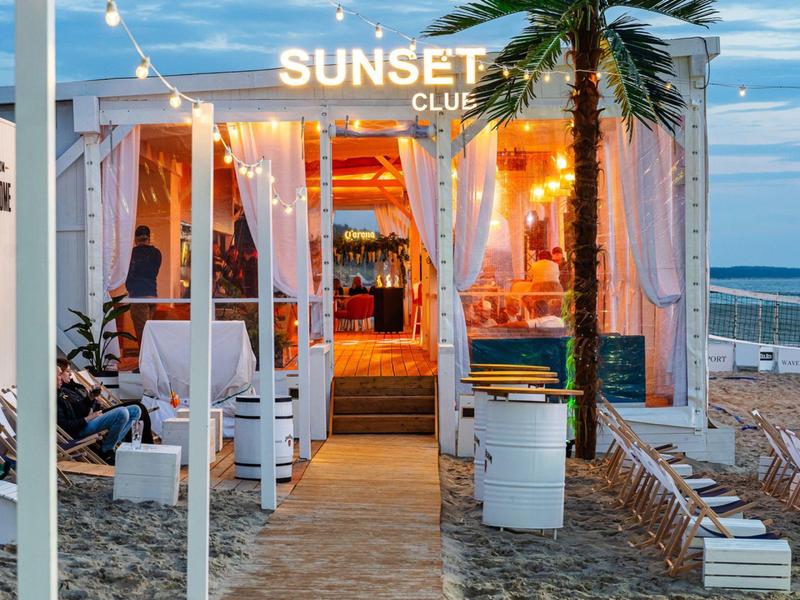 Strandbar met warm licht, witte gordijnen en planten op een zanderig pad bij zonsondergang.