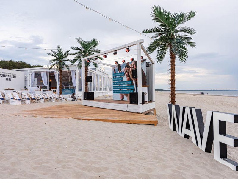 Strandbar met palmbomen, zand en een groot WAVE-bord naast een houten terras.