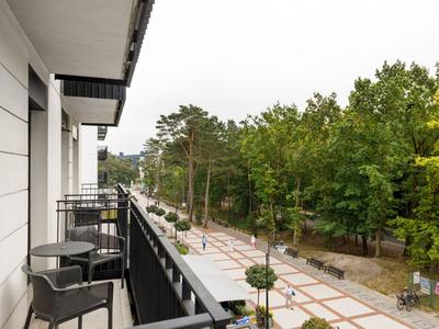 Balkon mit Stühlen und Tisch an Hotel mit Blick auf Baumallee und Gehweg.