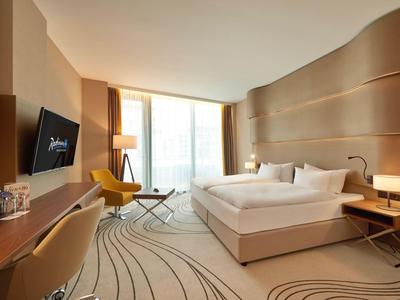 Modernes Hotelzimmer mit Doppelbett, Schreibtisch, Fernseher und großem Fenster mit Vorhängen.