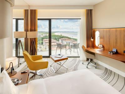 Modernes Hotelzimmer mit großem Bett, gelbem Sessel, Schreibtisch und Blick auf Balkon mit Landschaft.