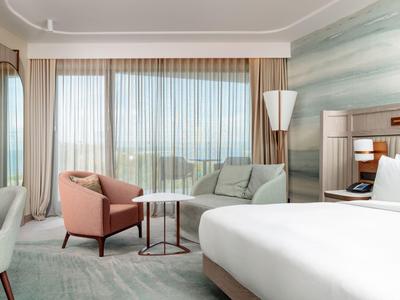 Helles Hotelzimmer mit großem Bett, zwei Sesseln, Tisch und weitem Fenster mit Vorhängen.