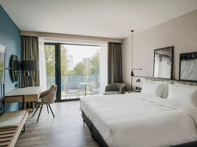 Modernes Hotelzimmer mit Doppelbett, Schreibtisch, Stuhl und Zugang zu einem Balkon mit Grünblick.