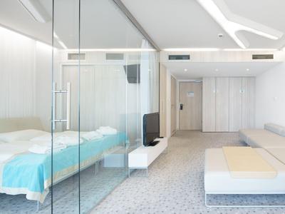 Modernes Hotelzimmer mit Glaswand, Bett, Sofa und weiterem TV-Bereich in hellem Design.