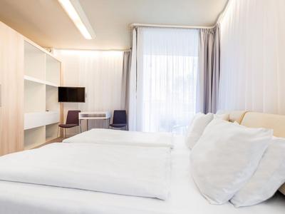 Helles Hotelzimmer mit großem Bett, weißen Vorhängen und moderner Einrichtung.