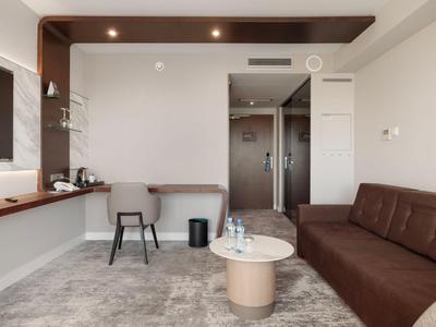 Modernes Hotelzimmer mit braunem Sofa, kleinem runden Tisch und Arbeitsbereich mit Stuhl.