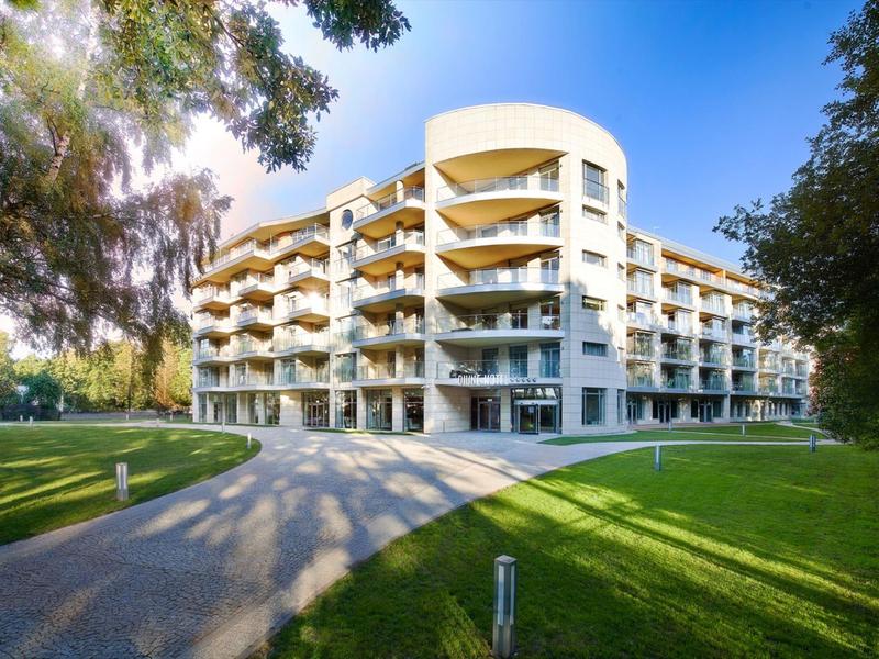 Edificio di hotel moderno con balconi, circondato da prati verdi e alberi sotto un cielo azzurro.