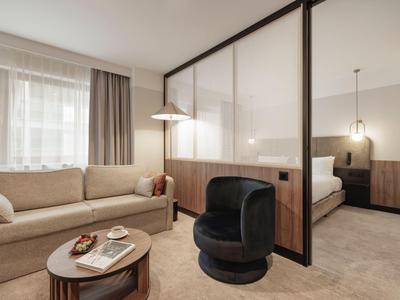 Modern eingerichtetes Hotelzimmer mit Couch, Sessel und durch Glaswand getrenntem Schlafzimmer.