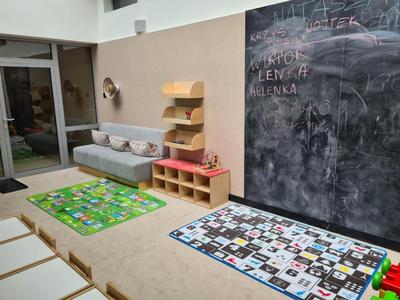 Area bimbi luminosa con cuscini, scaffali, due tappeti colorati e grande lavagna.