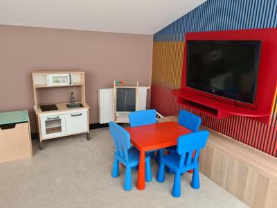 Camera per bambini con tavolo da gioco colorato, quattro sedie blu e cucina giocattolo accanto alla TV.