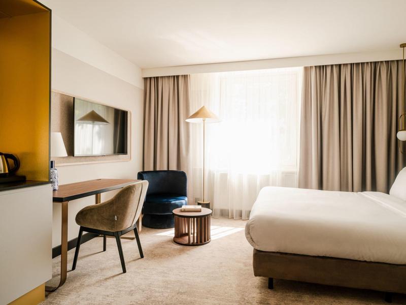 Camera d'hotel luminosa con grande finestra, letto, scrivania e poltrone in tonalità neutre.