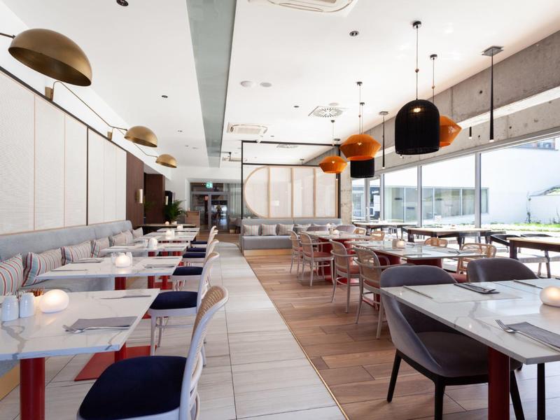 Ristorante luminoso con mobili moderni, colori legno e bianco, grandi finestre e vista esterna.