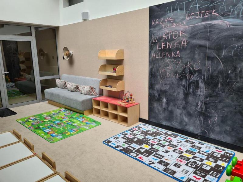 Area bimbi luminosa con cuscini, scaffali, due tappeti colorati e grande lavagna.