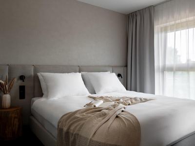 Camera d'hotel moderna con letto grande, cuscini bianchi e coperta beige vicino a una finestra con tende.