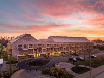 Großes Hotelgebäude bei Sonnenuntergang mit Parkplatz und begrüntem Vorplatz.