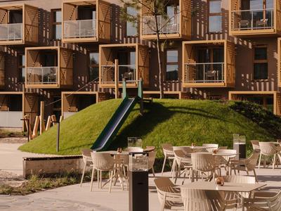 Moderner Hotelkomplex mit Holzstruktur, Balkonen und Outdoor-Sitzbereich bei Sonnenschein.