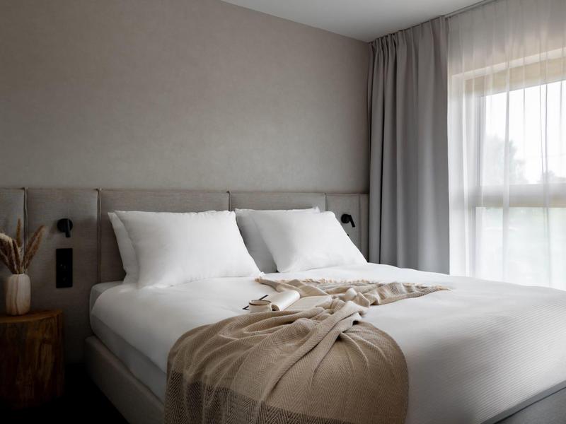 Camera d'hotel moderna con letto grande, cuscini bianchi e coperta beige vicino a una finestra con tende.