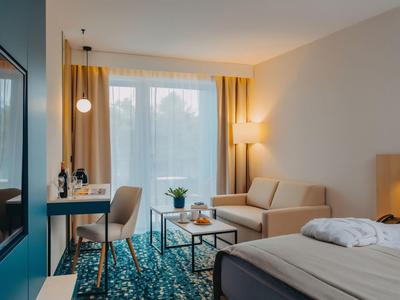 Camera d'albergo luminosa con letto, divano, scrivania e grande finestra con tende gialle.