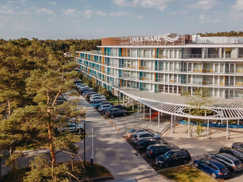 Hotel moderno su più piani accanto a una foresta con parcheggio pieno di auto.