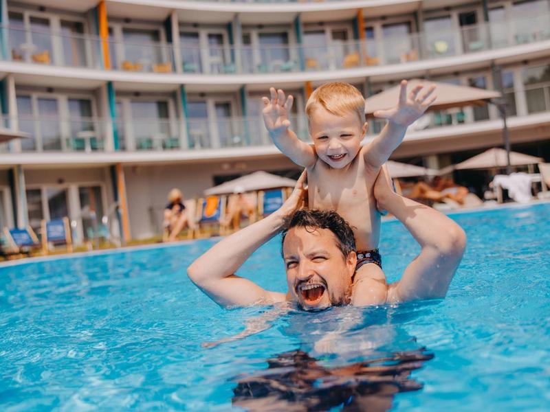 Padre e bambino ridono giocando nella piscina dell'hotel con architettura moderna sullo sfondo.