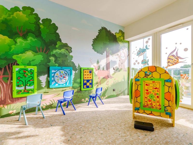 Sala giochi colorata per bambini con murales, pannelli di gioco e sedie vicino a grandi finestre.