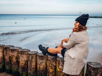 Žena v zimním oblečení sedí na dřevěných vlnolamech na pláži a dívá se na moře.