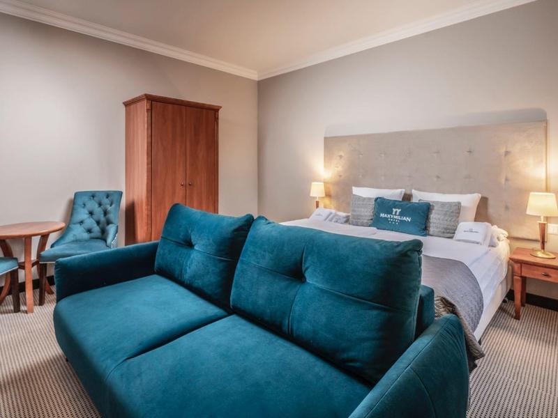 Moderní hotelový pokoj s manželskou postelí, modrou pohovkou a posezením za teplého světla.