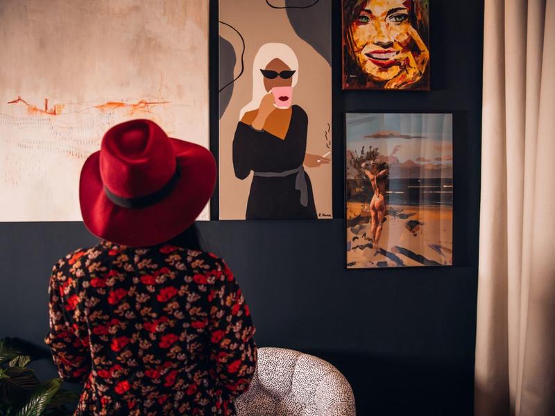 Osoba s červeným kloboukem se dívá na umělecká díla na tmavé stěně v hotelovém pokoji.