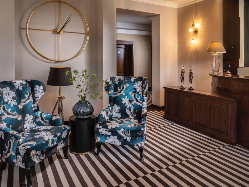 Elegantní hotelová hala s modrými vzorovanými křesly, pruhovanou podlahou a velkými zlatými nástěnnými hodinami.