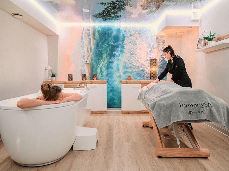 Persona in una vasca idromassaggio mentre un'altra riceve un massaggio in una stanza spa tranquilla.