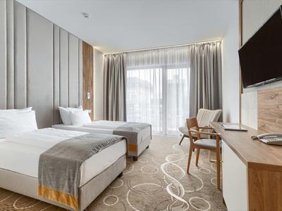 Camera d'albergo moderna con due letti singoli, sedie, scrivania e ampia finestra con tende.