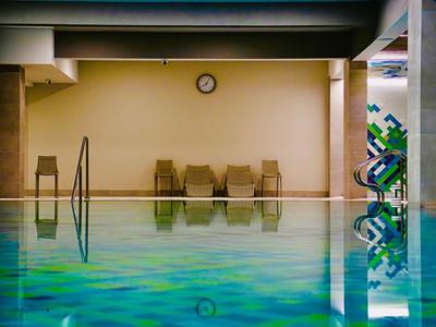 Piscina interna con sdraio e un orologio a muro sotto un soffitto con travi visibili.