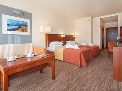 Pokój hotelowy z dwoma pojedynczymi łóżkami, sofą, stolikiem kawowym i telewizorem w ciepłych kolorach.