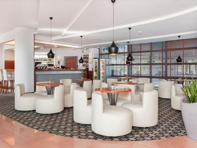 Área moderna de salón de hotel con sillones blancos, mesas estratégicamente ubicadas y lámparas colgantes.