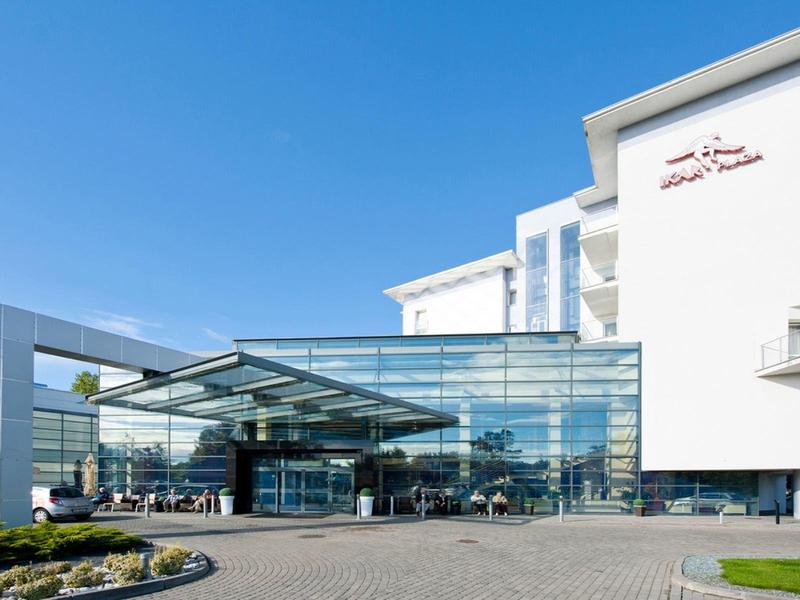 Nowoczesny hotel z dużą szklaną elewacją i tarasem na zewnątrz pod jasnym, błękitnym niebem.