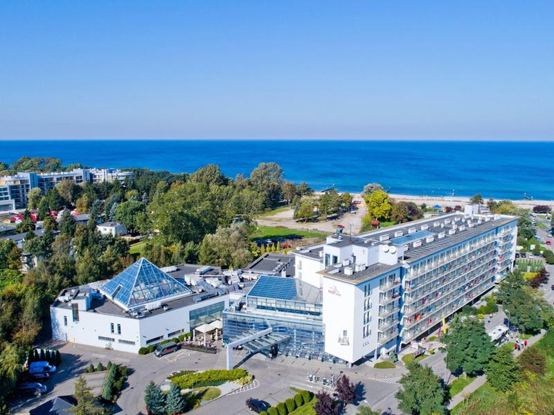 Duży budynek hotelowy na wybrzeżu z widokiem na niebieskie morze i otoczony drzewami.