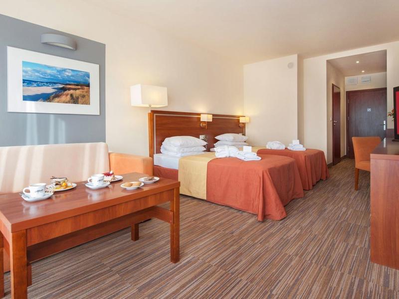 Pokój hotelowy z dwoma pojedynczymi łóżkami, sofą, stolikiem kawowym i telewizorem w ciepłych kolorach.