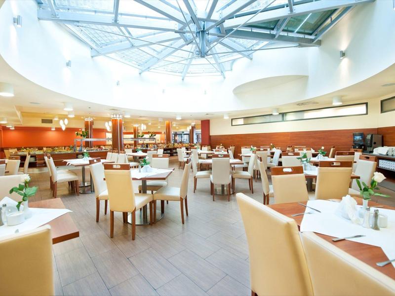 Duża przestrzeń restauracyjna hotelu z beżowymi krzesłami i okrągłym szklanym dachem.